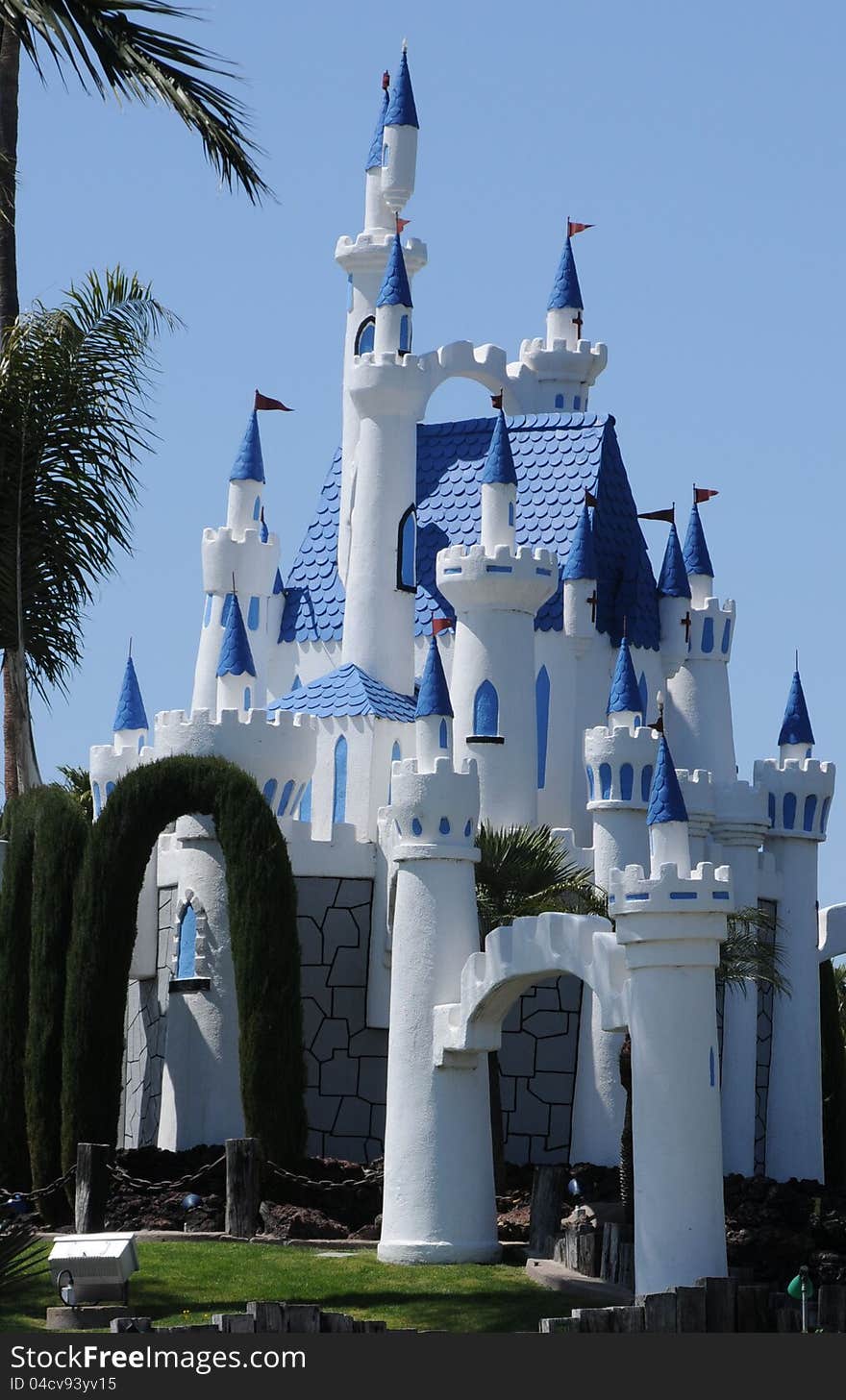 A fantasy castle in bright white wth blue trim, a brillant blue sky, green palm tree and some hedges border it