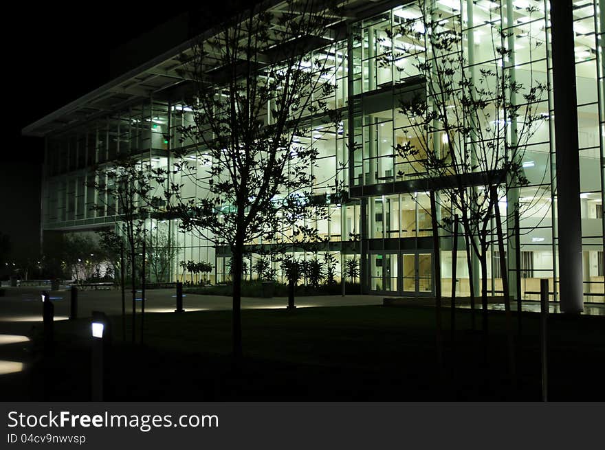 Evening view of a well lit glass and metal building with the dark shapes of lacey tree leaves and accent lights on the surrounding walkways. Evening view of a well lit glass and metal building with the dark shapes of lacey tree leaves and accent lights on the surrounding walkways