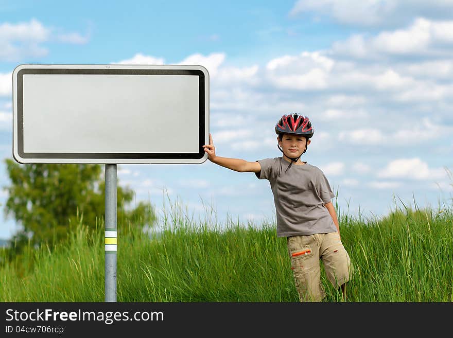 Boy cyclist leaning on blank sign with blue sky and green grass