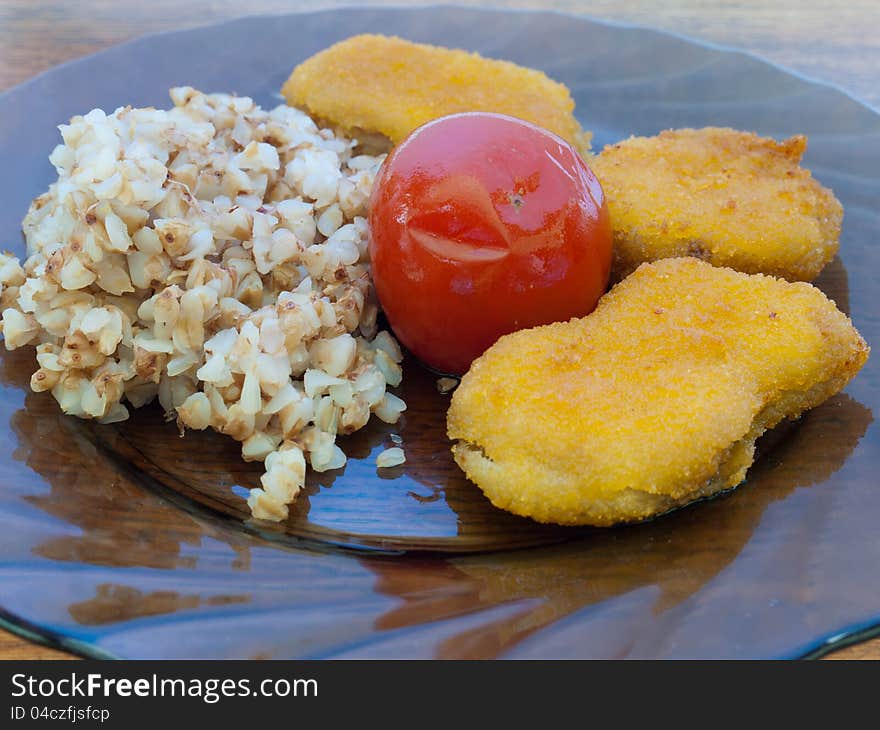 A plate of buckwheat porridge with chicken nuggets and tomato standing on a wooden table for the dinner.