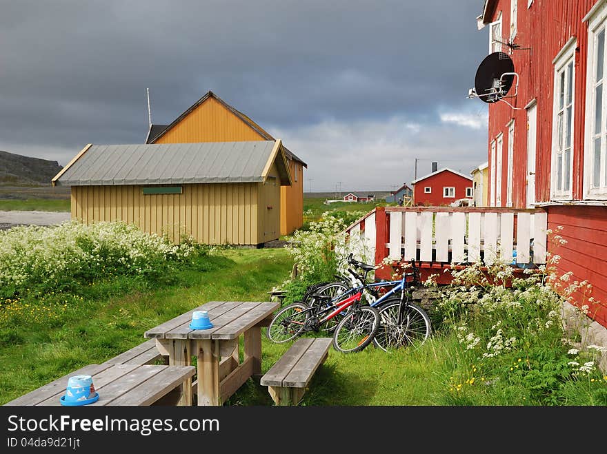 Norwegian fishing village is photographed in summer. Hamningberg is the easternmost point of Europe. There are several wood houses in the middle of the green grass under the overcast sky. Two bicycles are leaned against a wooden porch of the local cafe. Norwegian fishing village is photographed in summer. Hamningberg is the easternmost point of Europe. There are several wood houses in the middle of the green grass under the overcast sky. Two bicycles are leaned against a wooden porch of the local cafe.