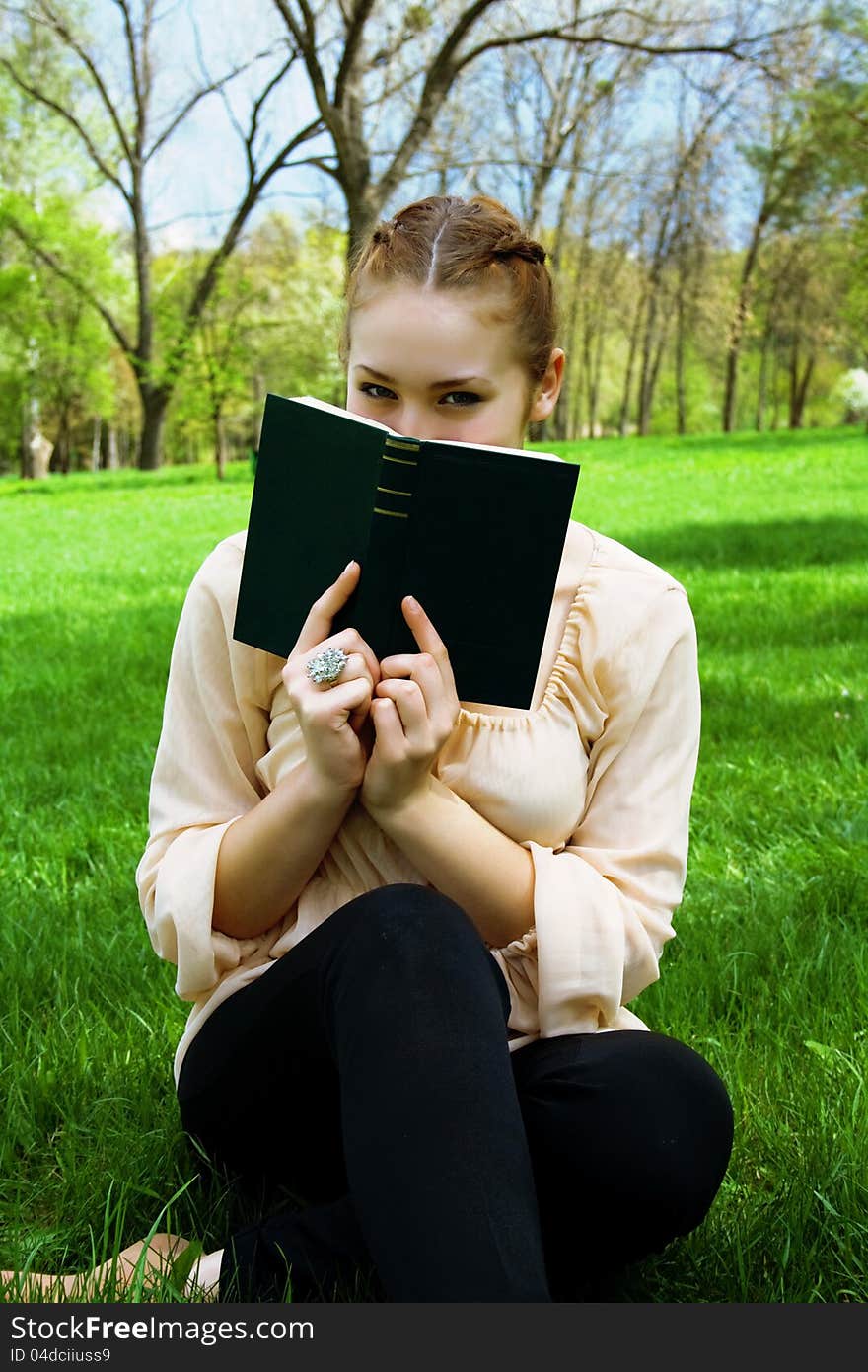 Woman hides her face behind a book sitting on green grass. Woman hides her face behind a book sitting on green grass