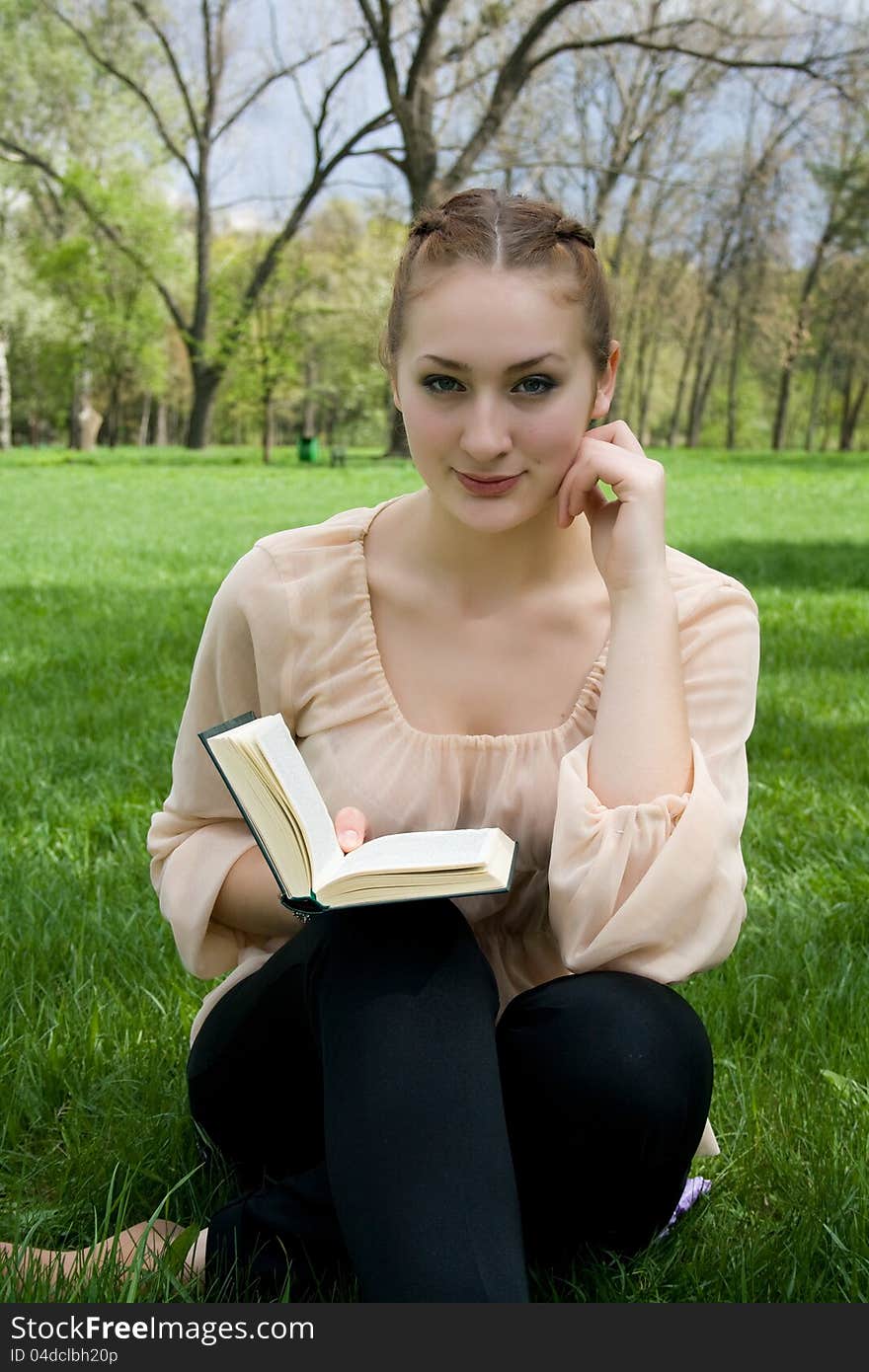 Young nice attentive woman lies on green grass and reads book against city park.