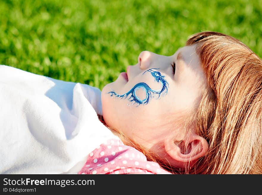 Portrait of a young girl with blue dragon on her face lying on a lawn