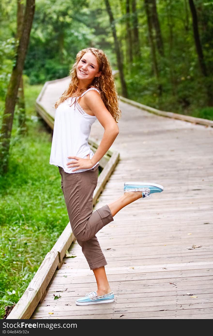 A beautiful blond teenage girl next door with curly hair wearing a white top having fun outdoors on a boardwalk in a local park. A beautiful blond teenage girl next door with curly hair wearing a white top having fun outdoors on a boardwalk in a local park.