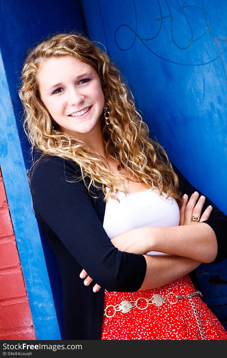 A beautiful blond teenage girl next door with curly hair and a pretty smile in a red skirt, white top, black sweater leaning against a blue door with her arms crossed in an urban setting. A beautiful blond teenage girl next door with curly hair and a pretty smile in a red skirt, white top, black sweater leaning against a blue door with her arms crossed in an urban setting.