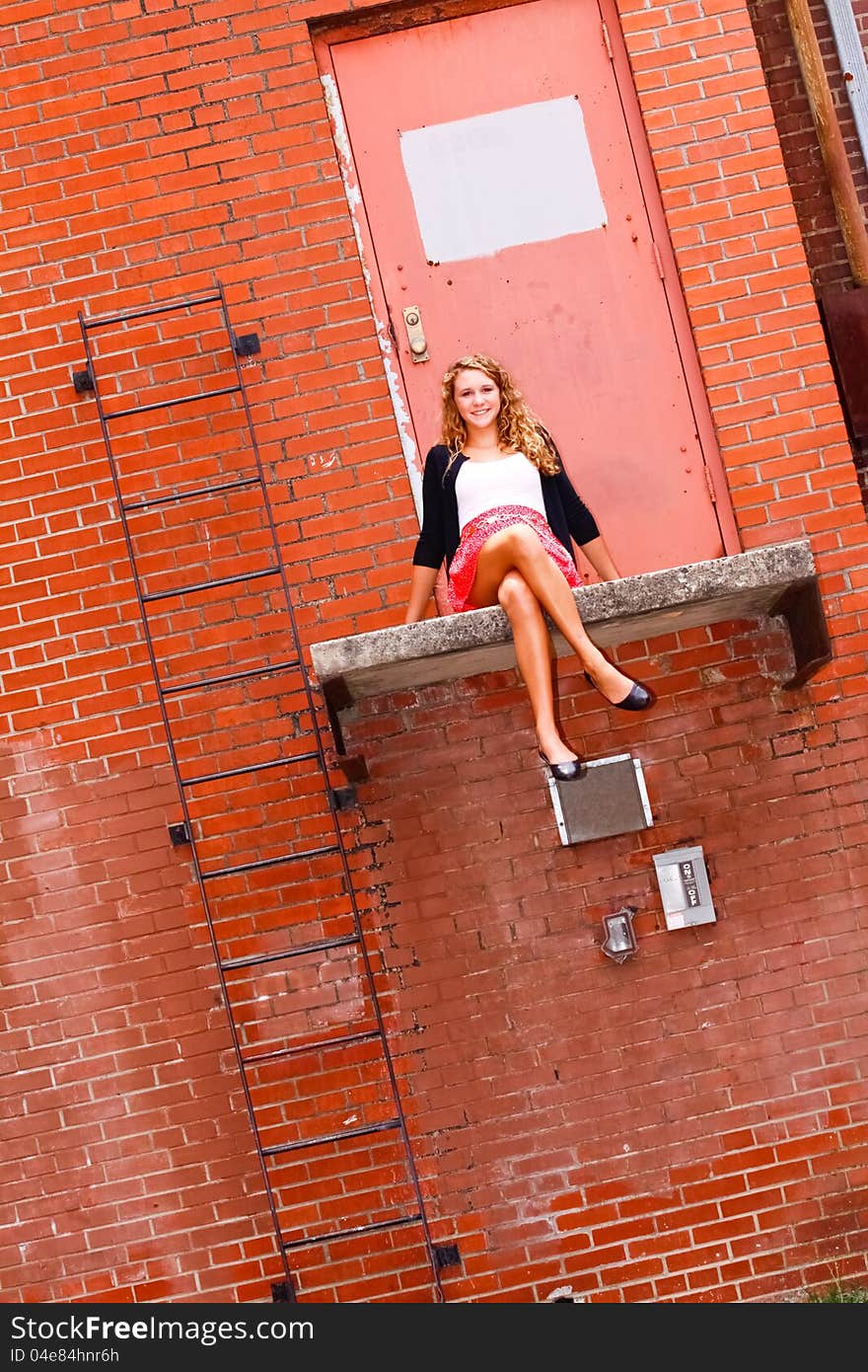 A beautiful blond teenage girl next door with curly hair and a pretty smile in a red skirt, white top, black sweater sitting on a ledge in front of a red brick wall and door in an urban setting. A beautiful blond teenage girl next door with curly hair and a pretty smile in a red skirt, white top, black sweater sitting on a ledge in front of a red brick wall and door in an urban setting.