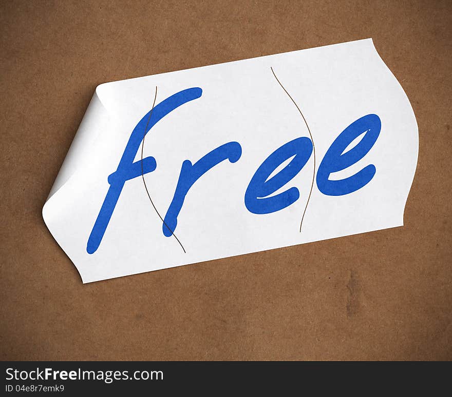 Free word hanwritten onto a tearable price tag over a cardboard background, blue color text, white label and brown carton. Free word hanwritten onto a tearable price tag over a cardboard background, blue color text, white label and brown carton