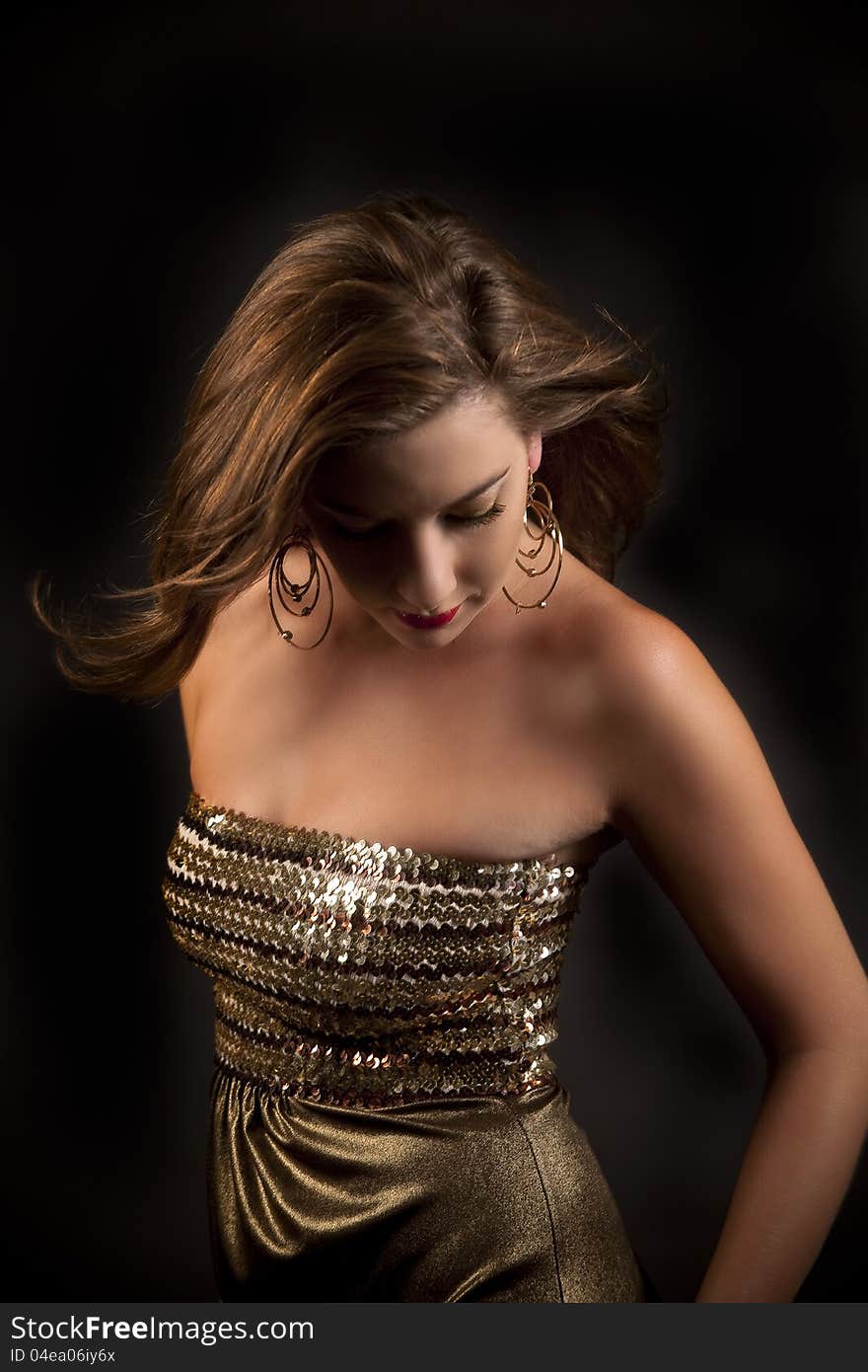 Beautiful caucasian woman in gold dress, gold earrings and dramatic makeup looking down with her hair blowing in the breeze against black background. Beautiful caucasian woman in gold dress, gold earrings and dramatic makeup looking down with her hair blowing in the breeze against black background.