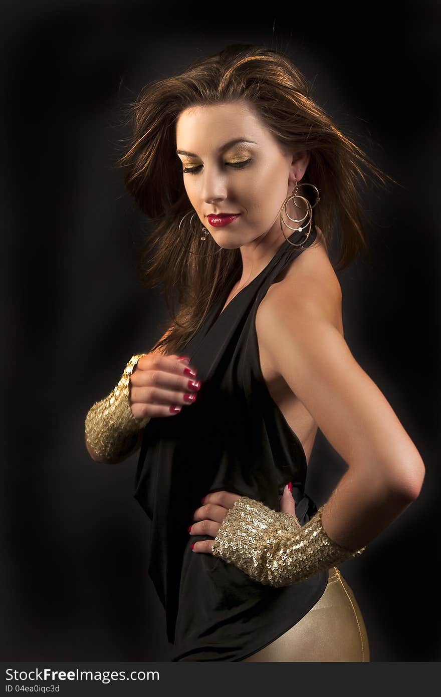 Beautiful woman with dramatic golden makeup, red lips, red nails, gold earrings and gloves looking down with her long hair blowing against black background. Beautiful woman with dramatic golden makeup, red lips, red nails, gold earrings and gloves looking down with her long hair blowing against black background