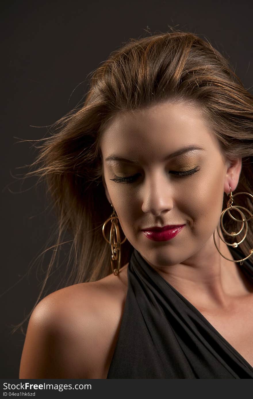 Beautiful caucasian woman in dramatic golden makeup with dark lashes and large golden drop earrings looking down while her long brown hair blows around her face against a dark background. Beautiful caucasian woman in dramatic golden makeup with dark lashes and large golden drop earrings looking down while her long brown hair blows around her face against a dark background