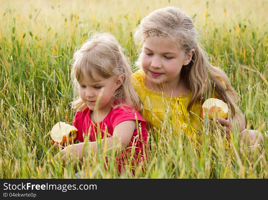 Two blonde girls sisters sitting in a wheat field and eating apples. Two blonde girls sisters sitting in a wheat field and eating apples