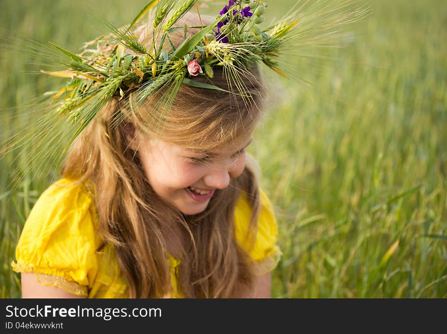 Beautiful little girl on the meadow. A wreath of wheat ears and wild flowers is on her blonde hair