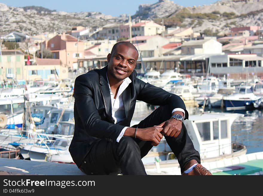 Young black man looking at camera and smiling, sitting somewhere at the seaside. A little port with some constructions and boats on background. Attractive, urban, positive, dynamic. Business wear. full-length portrait, outdoor, day light. Young black man looking at camera and smiling, sitting somewhere at the seaside. A little port with some constructions and boats on background. Attractive, urban, positive, dynamic. Business wear. full-length portrait, outdoor, day light.