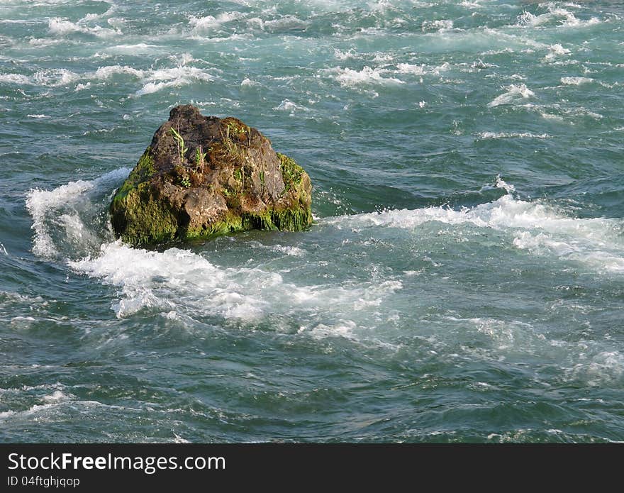 Single rock in the rapids of a fast flowing river with white water and strong current moving around it.