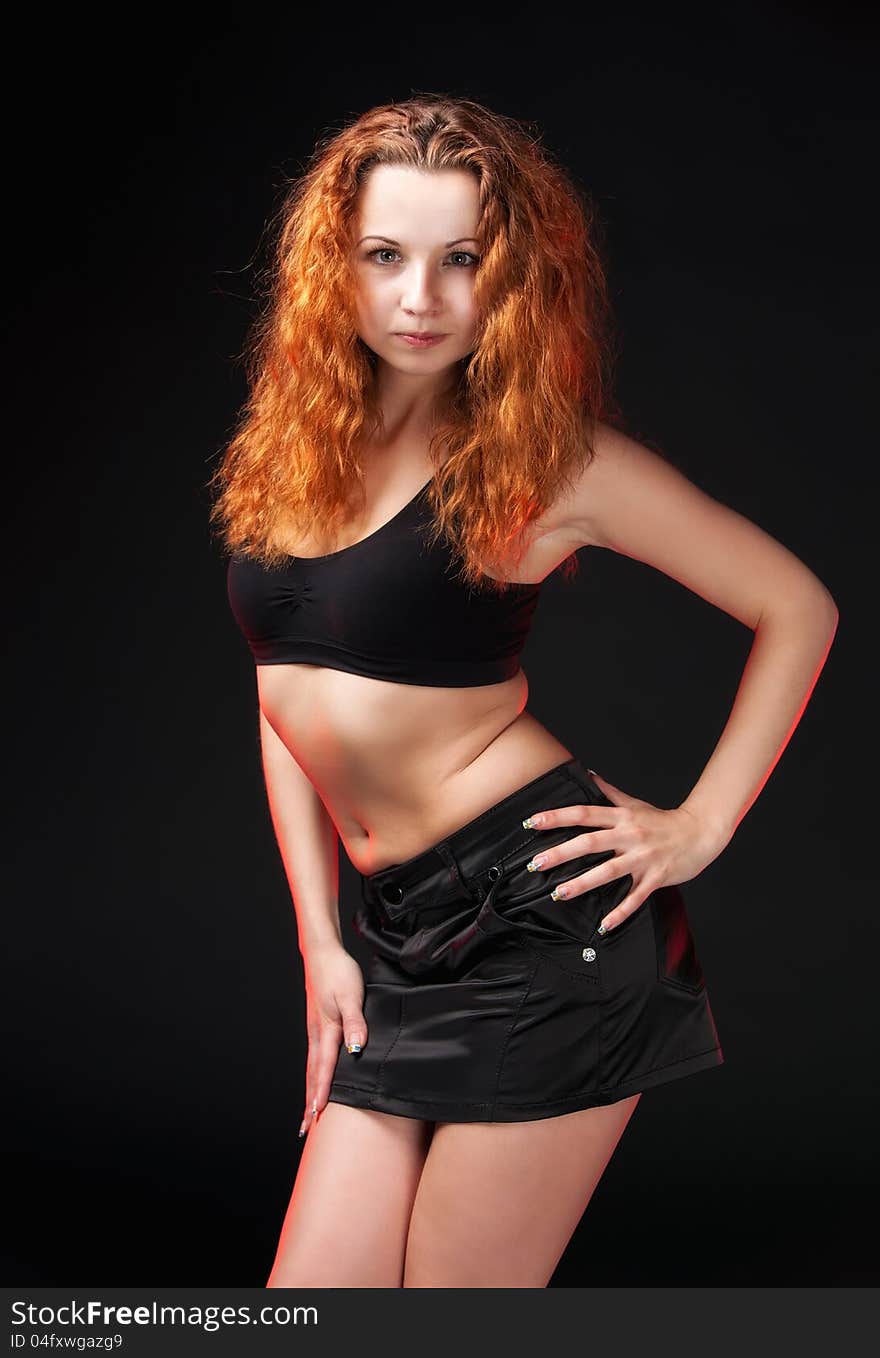 Portrait of a sexual redhead girl in a black bra and black leather skirt on a dark background