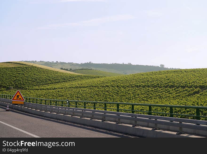 Warning sign of works on a highway, passing throw a small mountain planted with vines.