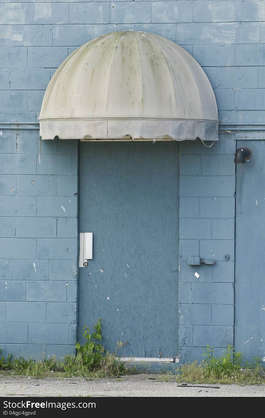 A blue metal door stand under a grungy canopy in an old blue cement block building. A blue metal door stand under a grungy canopy in an old blue cement block building