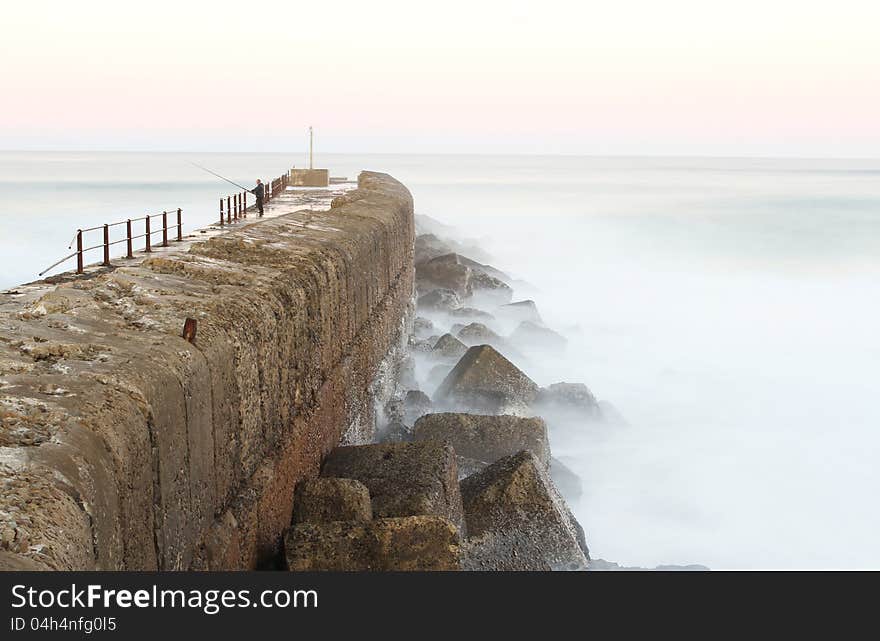 One solitary man is standing on a peer fishing just after sunset in Port Alfred, South Africa. It is a long exposure. One solitary man is standing on a peer fishing just after sunset in Port Alfred, South Africa. It is a long exposure.