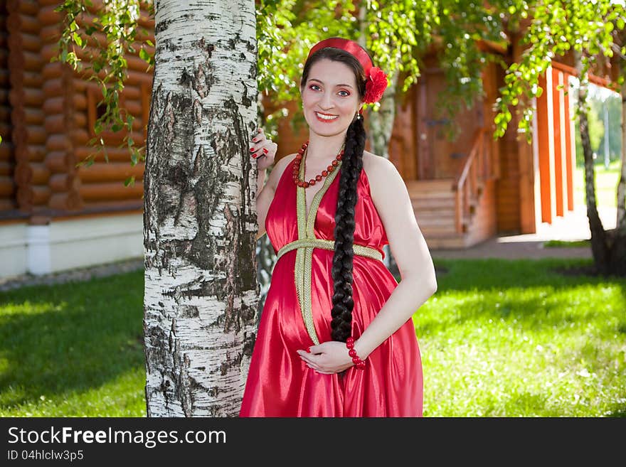 A happy young pregnant girl is standing under a tree in a red dress