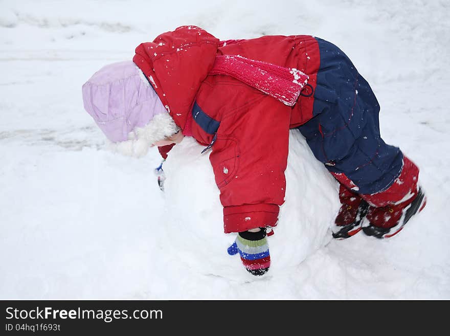 Little girl wearing warm jumpsuit lies on big snowball; child rolls snowball. Little girl wearing warm jumpsuit lies on big snowball; child rolls snowball