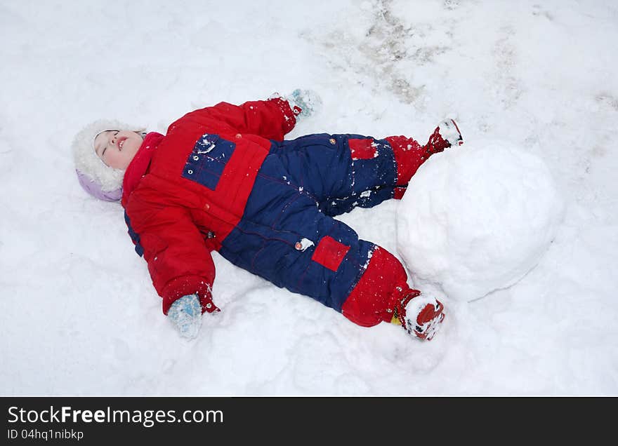 Little tired girl wearing warm jumpsuit lies on snow near big snowball. Little tired girl wearing warm jumpsuit lies on snow near big snowball