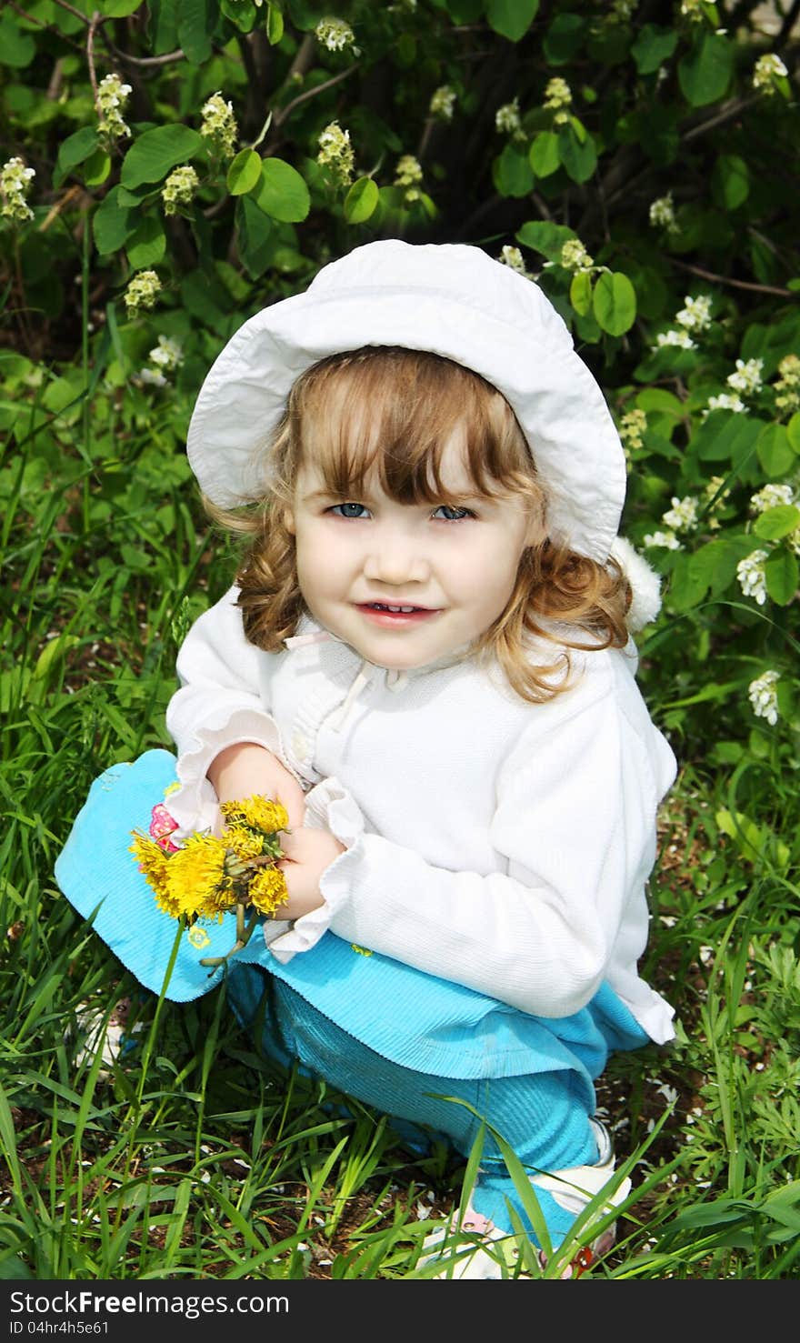 Beautiful little girl wearing white panama holds yellow dandelions and sits on grass. Beautiful little girl wearing white panama holds yellow dandelions and sits on grass