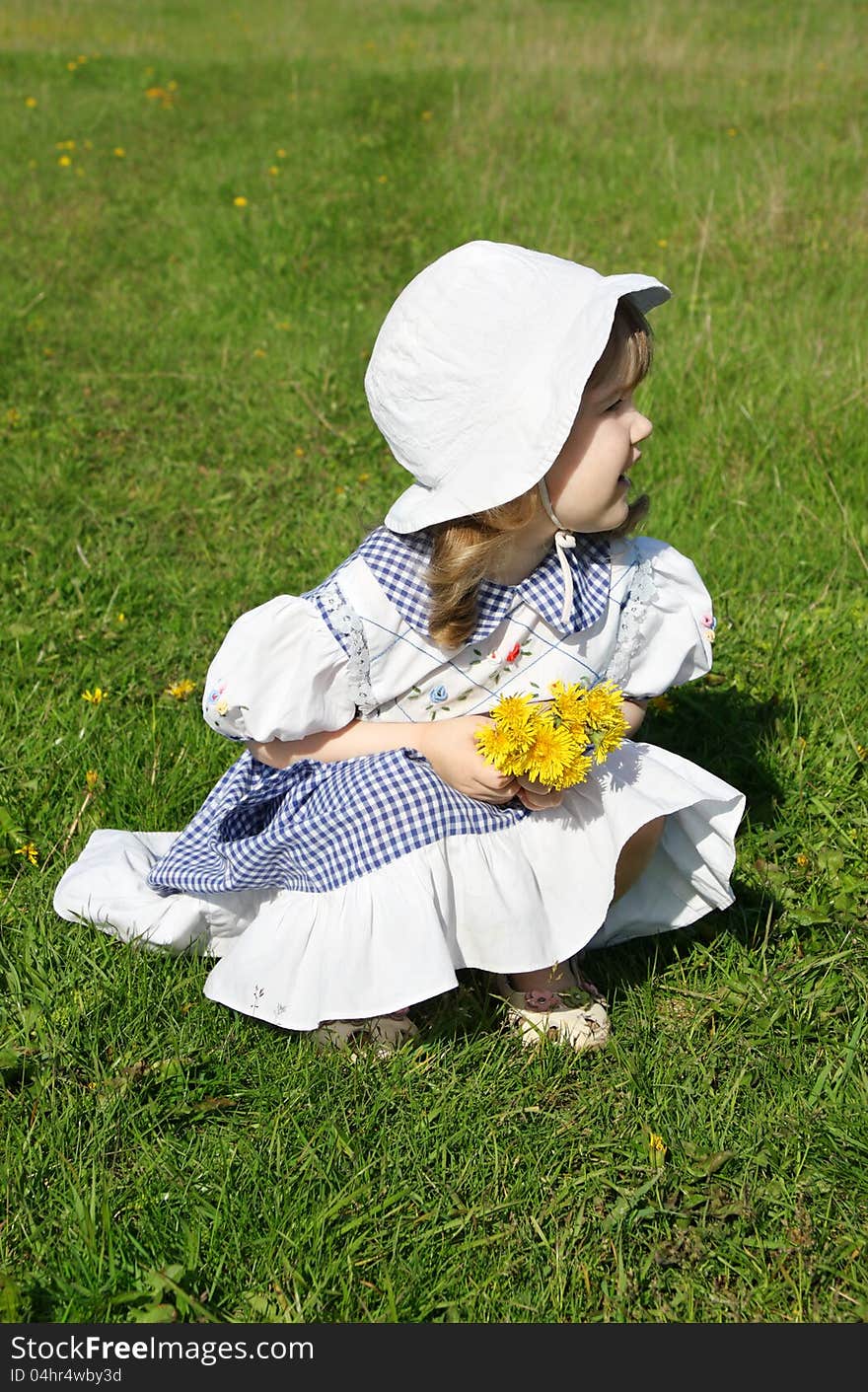 Beautiful little girl wearing dress with yellow dandelions sits on grass and looks into distance. Beautiful little girl wearing dress with yellow dandelions sits on grass and looks into distance