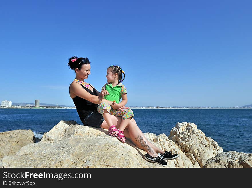 Mother and daughter sitting on his knees and laughing on the cliff by the sea. Mother and daughter sitting on his knees and laughing on the cliff by the sea