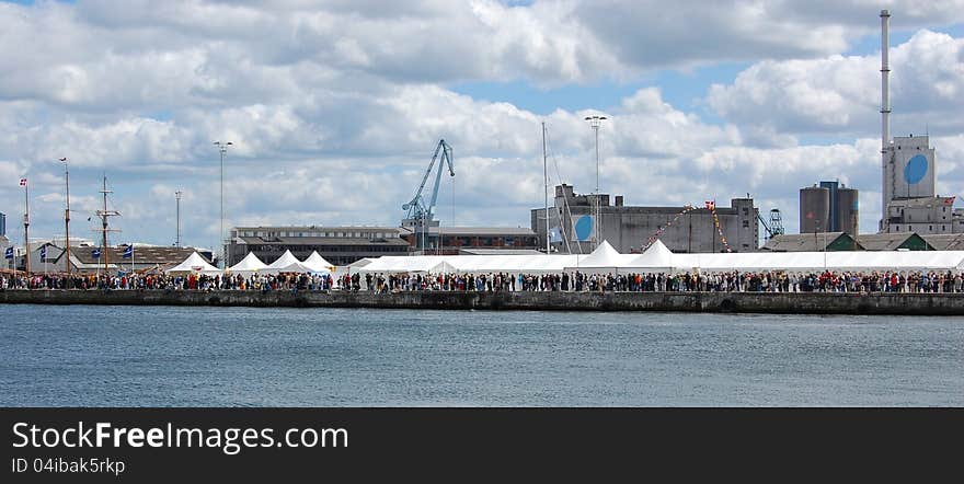 Crowds at a sailing festival in Aarhus, Denmark, gather along and in the white tents placed on the industrial harbor quay with silos and cranes. Crowds at a sailing festival in Aarhus, Denmark, gather along and in the white tents placed on the industrial harbor quay with silos and cranes.