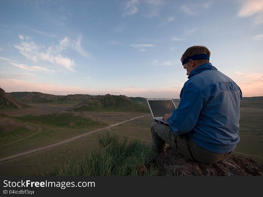 The man with the laptop sits on a hill against the evening sky