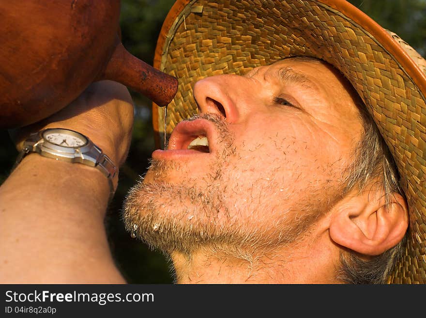 Horizontal image made on sweltering late afternoon to a man drinking water from a jug. He is beardy, seems to be thirsty and tired, and wears a straw hut. Horizontal image made on sweltering late afternoon to a man drinking water from a jug. He is beardy, seems to be thirsty and tired, and wears a straw hut.