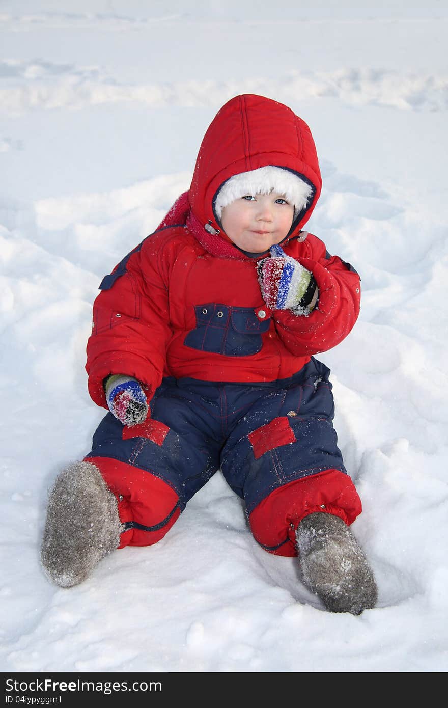 Little girl wearing red jumpsuit sits on snow and eats snow at winter. Little girl wearing red jumpsuit sits on snow and eats snow at winter