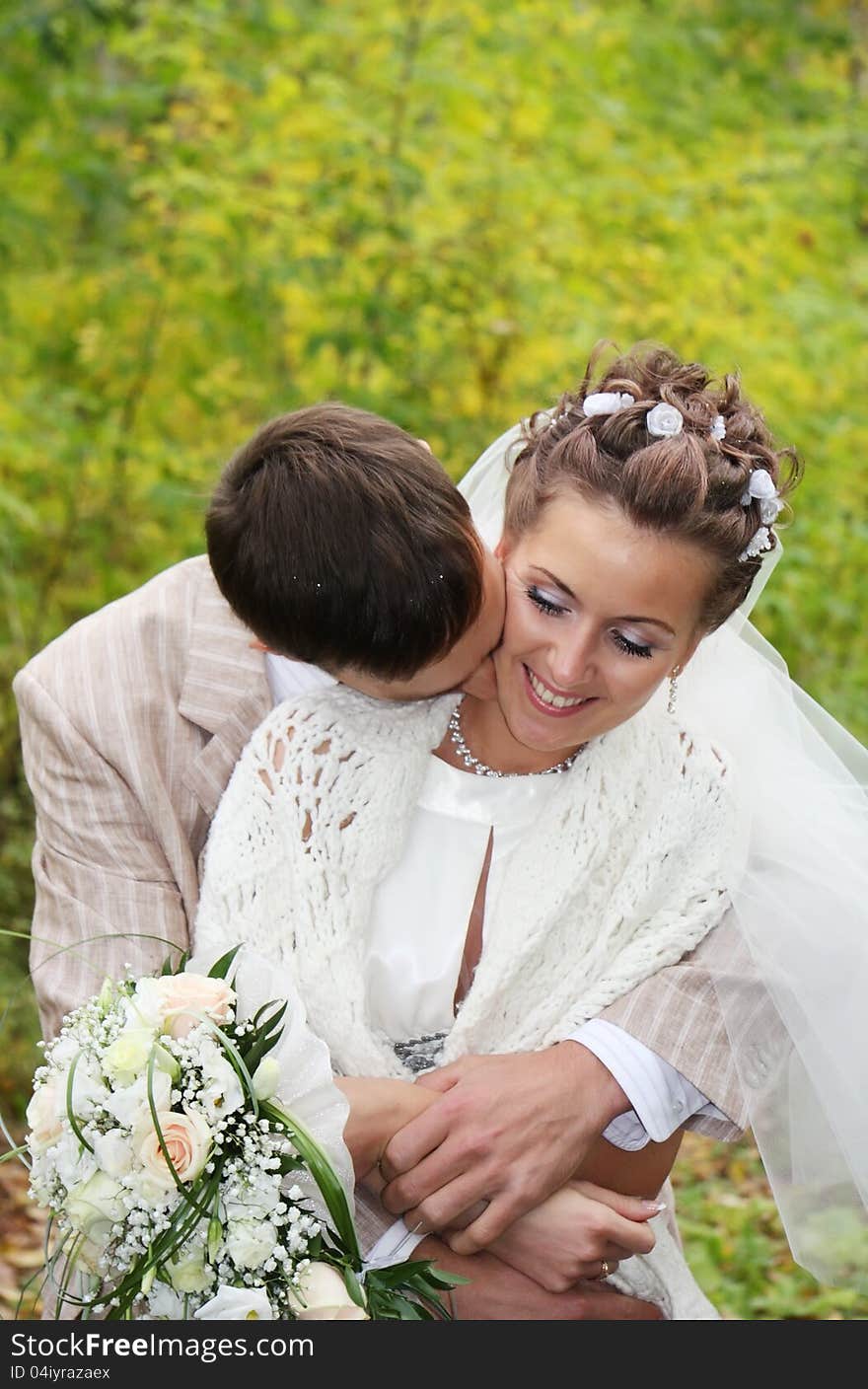 Groom hugs his beautiful bride with roses and kisses neck in autumn park. Groom hugs his beautiful bride with roses and kisses neck in autumn park