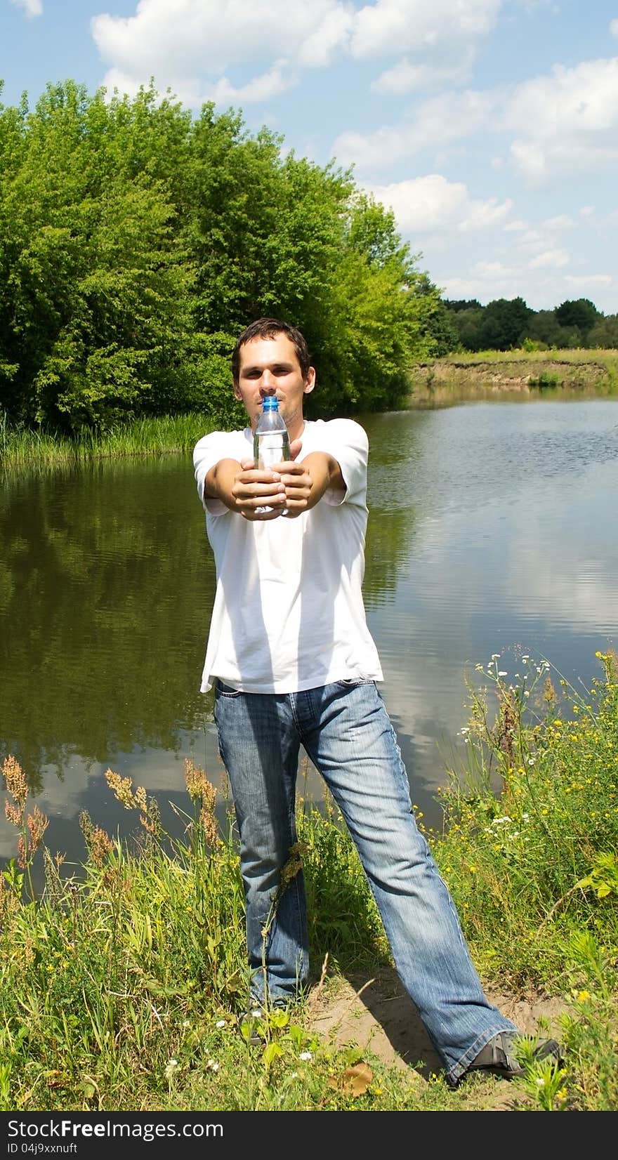 Young man stands near the river holding in his outstretched hands a bottle of water