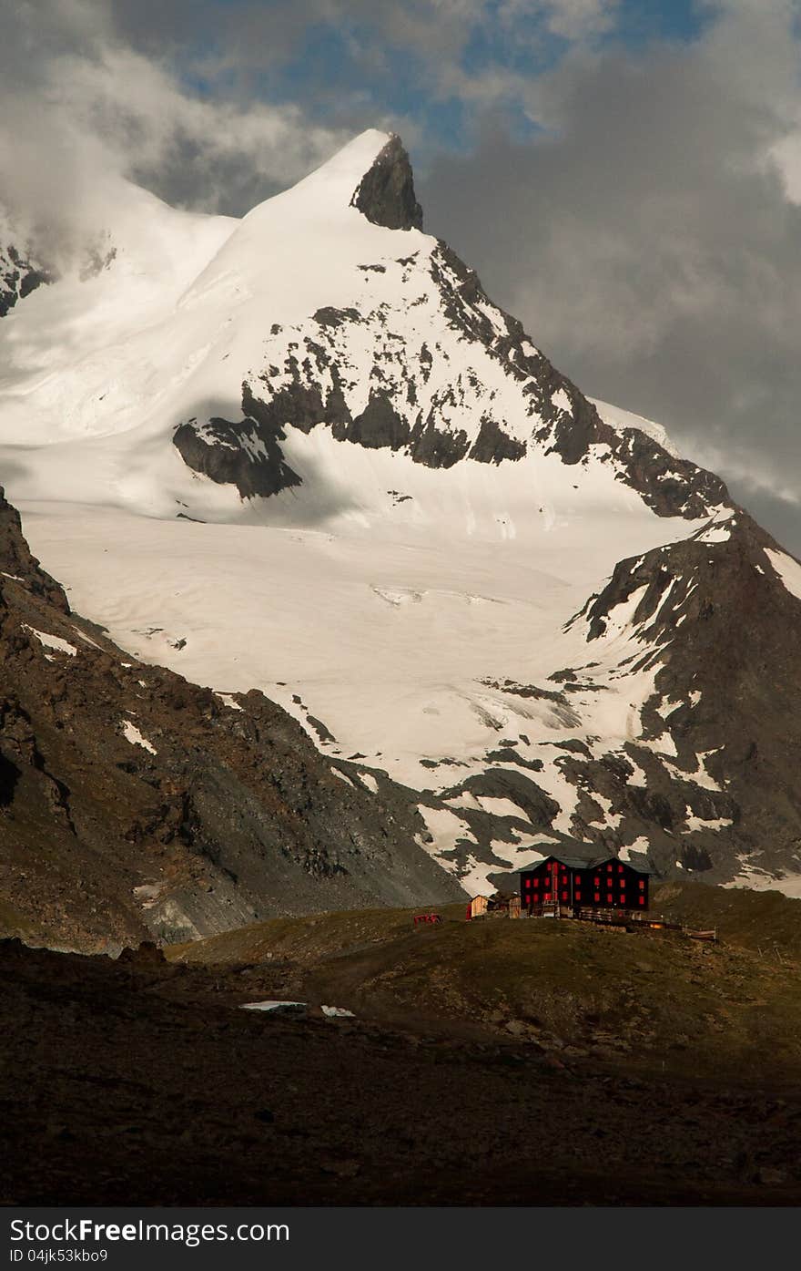 Fluhalp mountain hut under the massive mountain peak with cloudy sky.