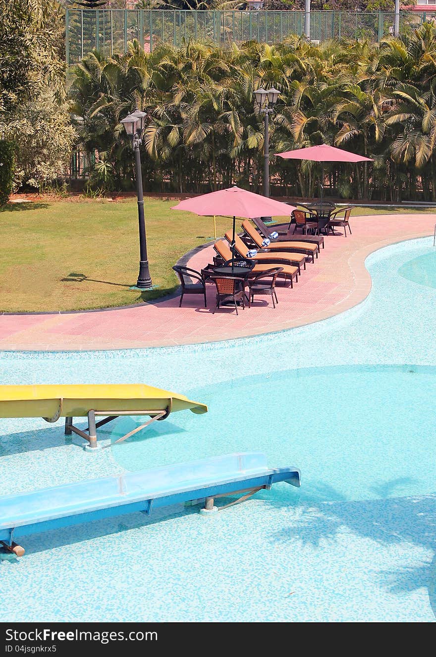 A beautiful swimming pool with clean blue water and relaxing chairs with bath towels, umbrella on poolside in a resort on a bright sunny summer day. A beautiful swimming pool with clean blue water and relaxing chairs with bath towels, umbrella on poolside in a resort on a bright sunny summer day.