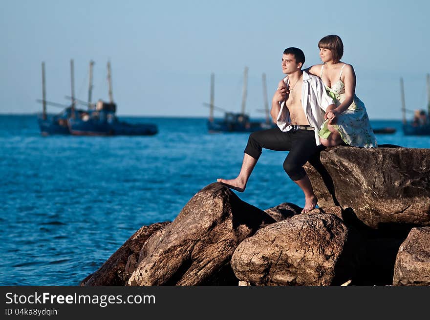 Young couple sitting on rocks near the lake on the background of sailing ships. Young couple sitting on rocks near the lake on the background of sailing ships