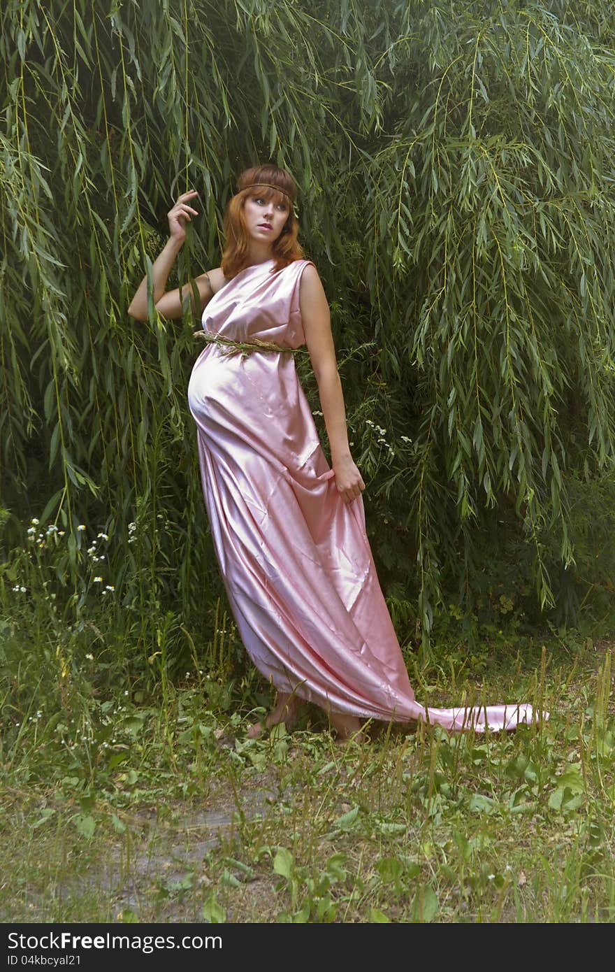 A pregnant woman in a beautiful pink long dress looks somewhere in the direction of hugging a tree branch. A pregnant woman in a beautiful pink long dress looks somewhere in the direction of hugging a tree branch.