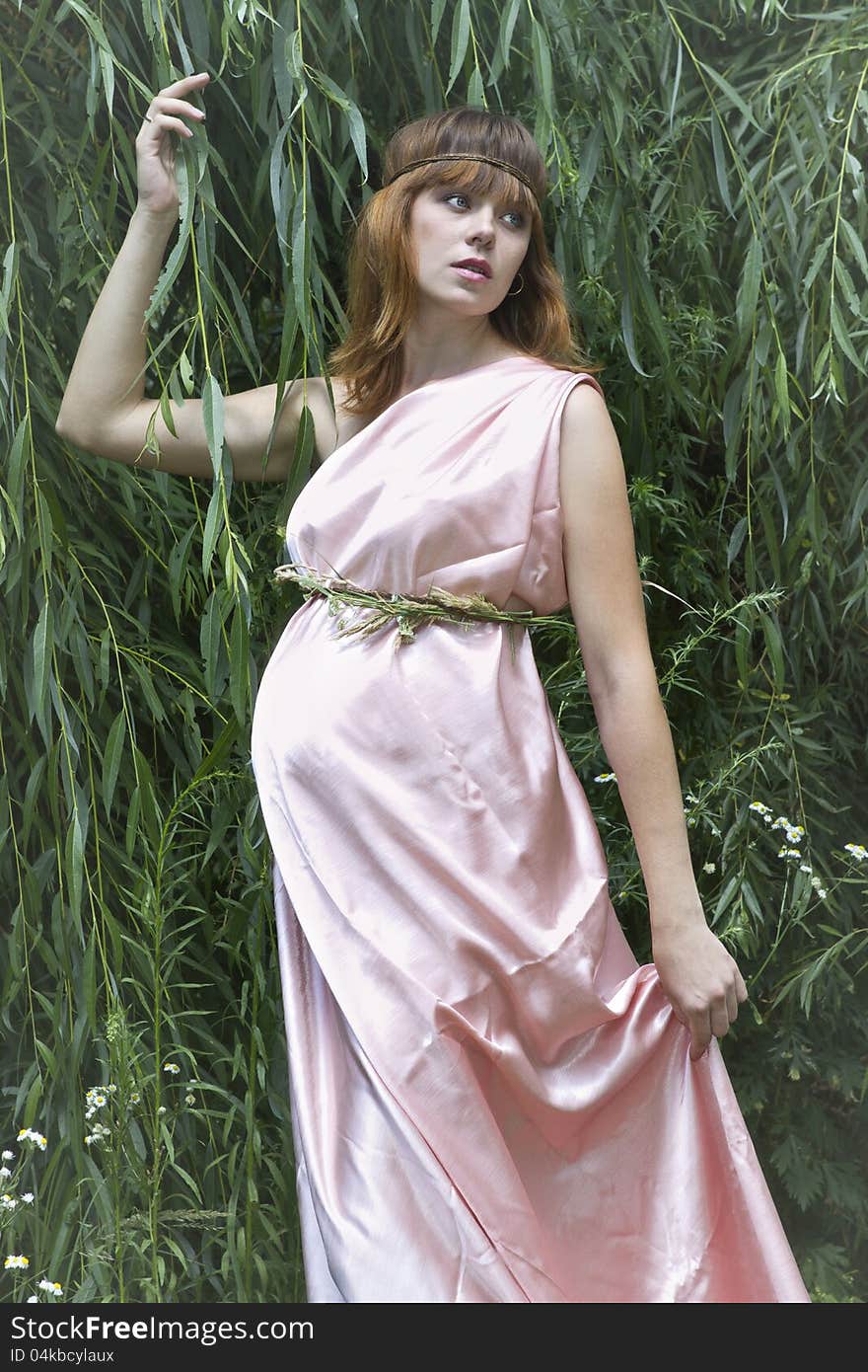 A pregnant woman in a beautiful pink long dress looks somewhere in the direction of hugging a tree branch. A pregnant woman in a beautiful pink long dress looks somewhere in the direction of hugging a tree branch.