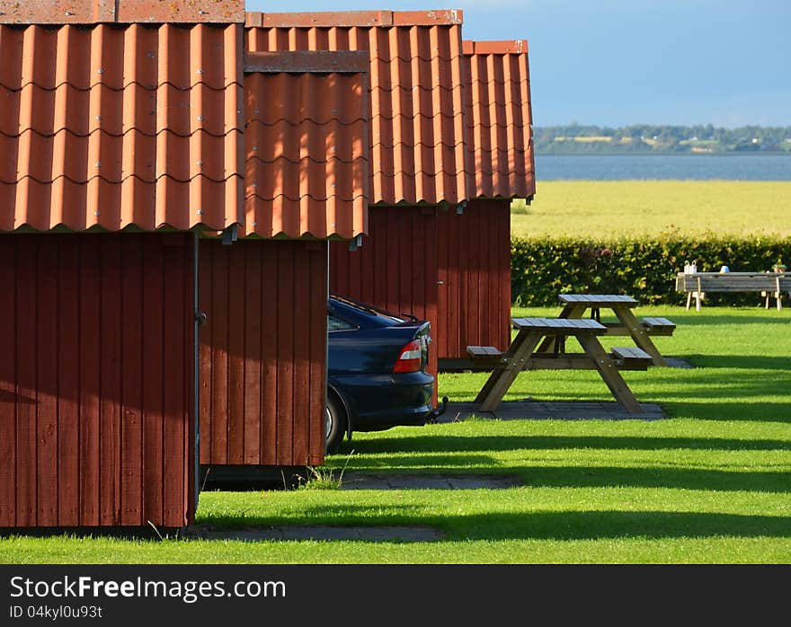 Four red holiday cottages with red tiled roofs form a line pointing to the bay behind a field. A car is parked between them and benches with tables are in front. The evening sun creates long shadows in the grass. Four red holiday cottages with red tiled roofs form a line pointing to the bay behind a field. A car is parked between them and benches with tables are in front. The evening sun creates long shadows in the grass.