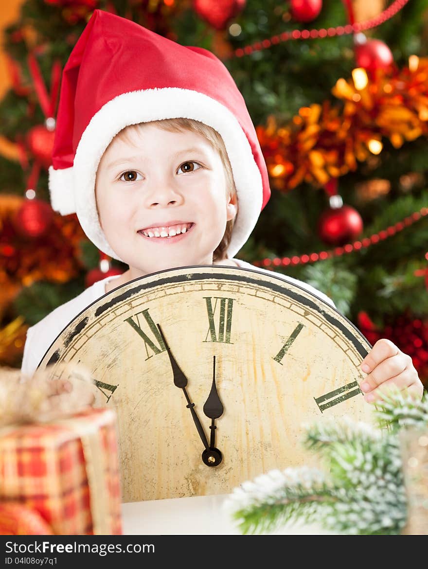 Happy kid in Santa`s hat holding old wooden clock against decorated Christmas background. Happy kid in Santa`s hat holding old wooden clock against decorated Christmas background