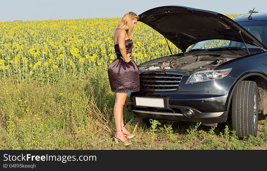 Bemused woman standing looking at the engine compartment of her car which has broken down alongside a field of sunflowers in the countryside. Bemused woman standing looking at the engine compartment of her car which has broken down alongside a field of sunflowers in the countryside