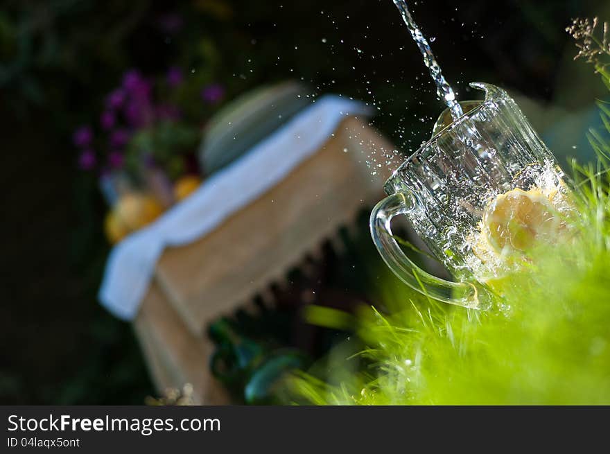 Sparkling water puring and splashing into lemon filled lemonade jug at picnic in the fresh green grass with picnic table laid out in the background. Sparkling water puring and splashing into lemon filled lemonade jug at picnic in the fresh green grass with picnic table laid out in the background