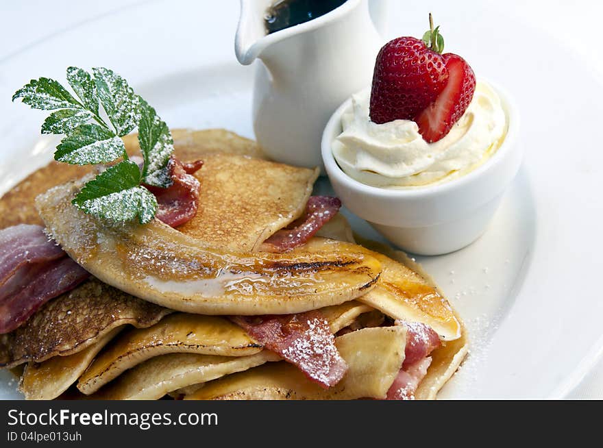 Stack of breakfast pancakes with bacon and banana. A jug of maple syrup and pot of cream accompanies it