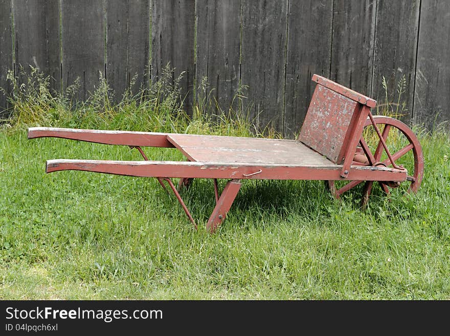 Very old wheel barrow made of wood and metal, painted red, sitting in a grassy field in front of an old wood fence. Very old wheel barrow made of wood and metal, painted red, sitting in a grassy field in front of an old wood fence.