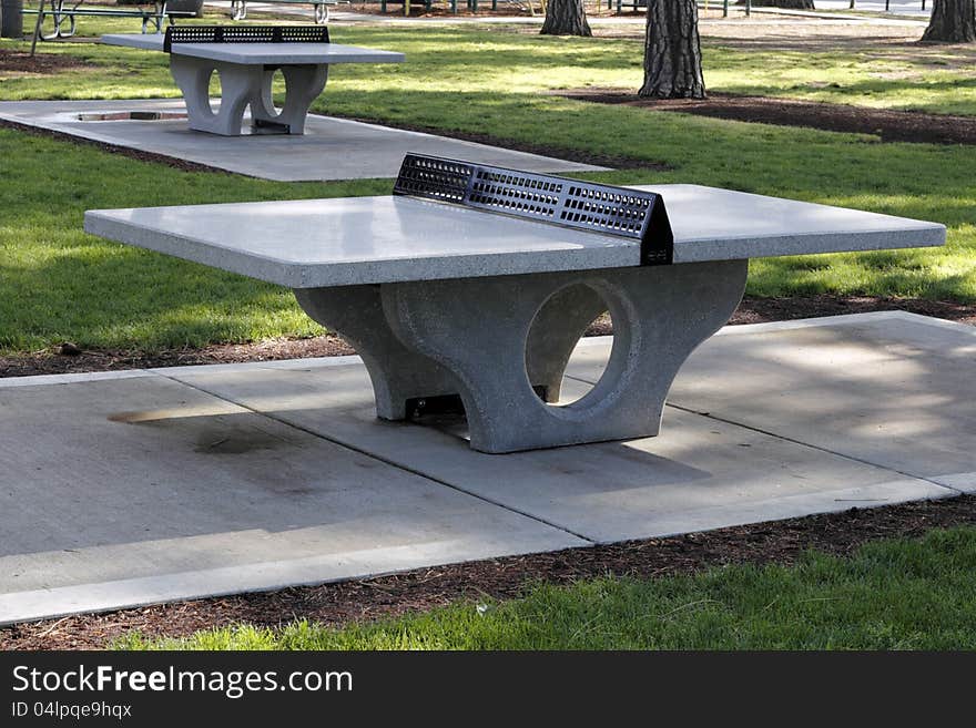 Outdoors in a city park are two cement ping pong tables surrounded by grass and trees in a mostly shady spot on a spring day.