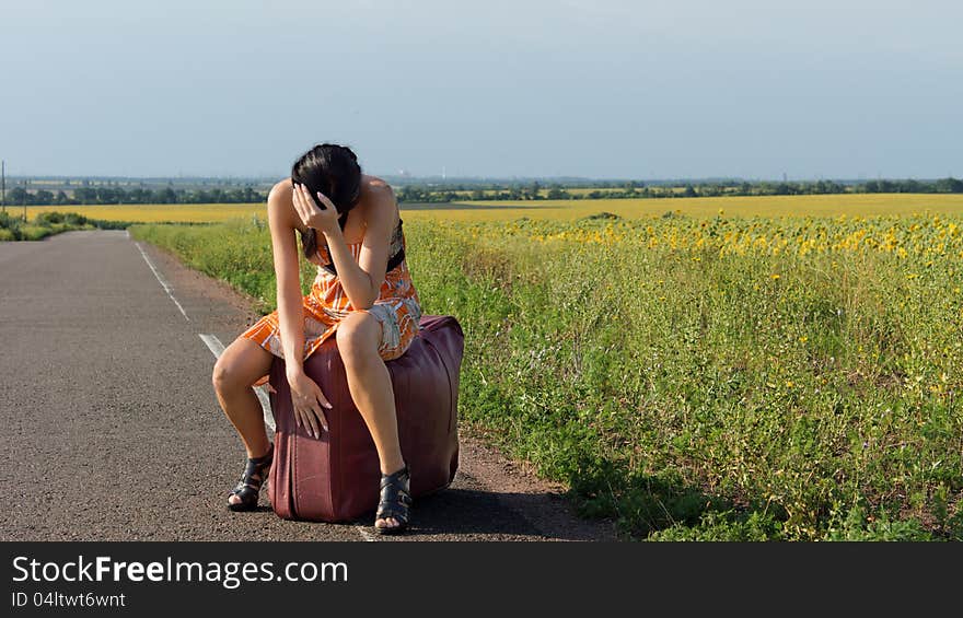 Despondent female hitchhiker in stilettoes and a sundress sitting on a large suitcase alongside a country road with her head in her hands