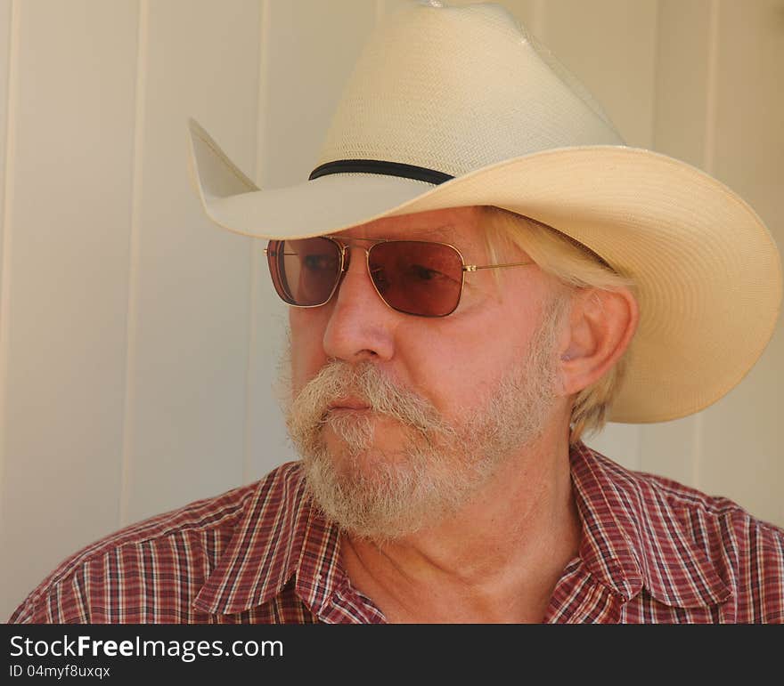 An older bearded man with a white cowboy hat, checked shirt and dark glasses is looking off to the side, with a serious concerned look. An older bearded man with a white cowboy hat, checked shirt and dark glasses is looking off to the side, with a serious concerned look