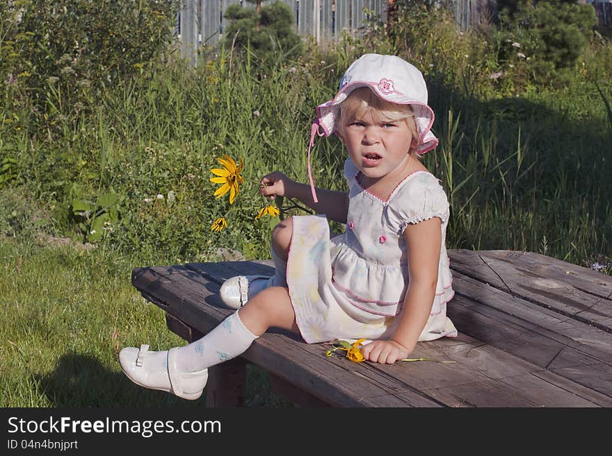 Happy child the girl in a white dress and a white cap with flower sitting on green grass outdoors in spring garden. Happy child the girl in a white dress and a white cap with flower sitting on green grass outdoors in spring garden